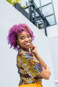 Kabrena Williams stands against a grey building, she is smiling while looking over her shoulder and is wearing a colorful print shirt