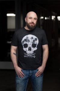 Dan Bennett wears a black shirt with skull and palm tree design. He is smiling at the camera and stands against a black background.