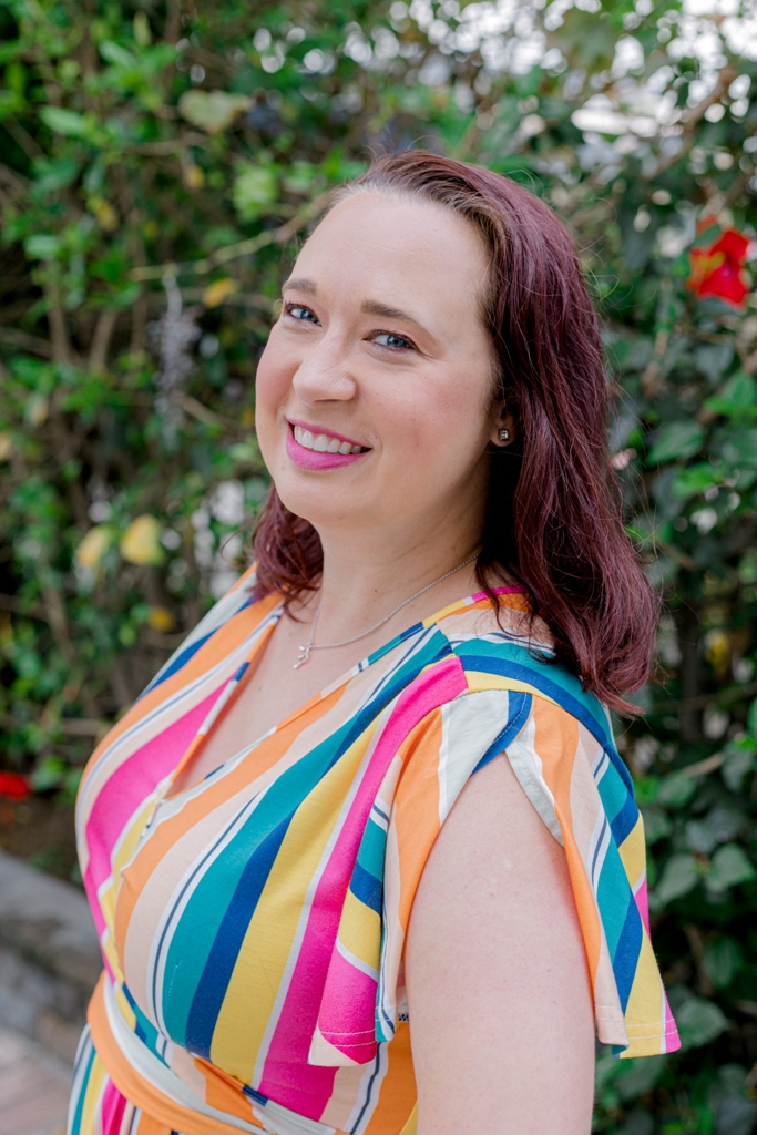 Mindy Marzec stands in front of greenery. She is looking over her shoulder and wears a rainbow stripe dress